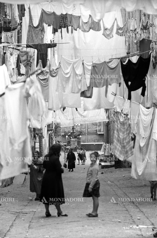 View of a sloping alley in the Old Town, the clothes, hanging on wires, occupy most of the sight; a child smiling and an elderly woman stand in the middle of the road on the foreground. Naples (Italy), May 1957. SPECIAL FEE  - CONTACT US FOR NOT EDITORIAL USAGE