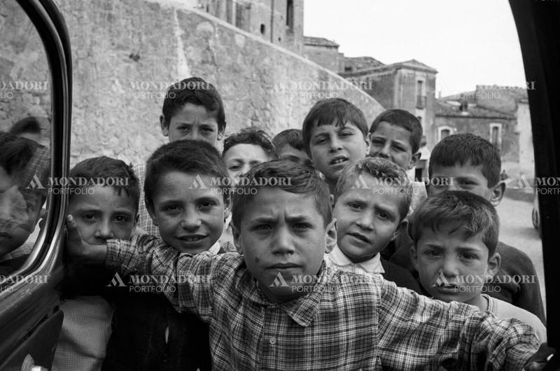 A bunch of kids are flocking one side to another to appear in the shot, near the walls of the rural village of Trebisacce; their faces show a mixture of excitement, curiosity and boldness. Trebisacce (Cosenza), Italy, May 1957. SPECIAL FEE  - CONTACT US FOR NOT EDITORIAL USAGE