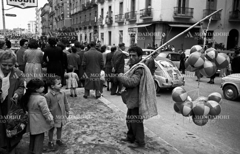 Along the street, two children look amazed a coster selling balls; behind them, a crowd of people follow a hearse in the distance. Naples (Italy), 1960. SPECIAL FEE  - CONTACT US FOR NOT EDITORIAL USAGE