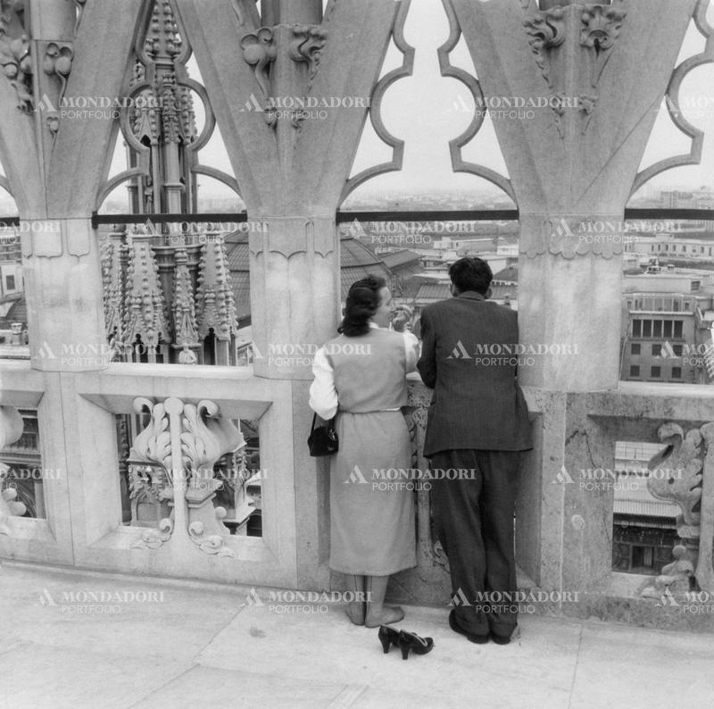 A couple during honeymoon, filmed from behind on the terraces of Milan Cathedral, leaning on the stone parapet under a gothic cusp; they admire the beautiful relief of one of the big spires of the roof, and the glass and iron dome of Galleria Vittorio Emanuele II in the background; she took off her heeled shoes to be comfortable during this cultural itinerary. Milan (Italy), 1955 SPECIAL FEE  - CONTACT US FOR NOT EDITORIAL USAGE