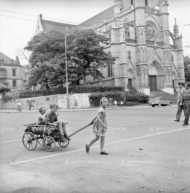 A little girl pulling a cart with two children on board in front of a church during the Geneva Summit to discuss global security, German unification and disarmament. Geneva, 18th July 1955 SPECIAL FEE  - CONTACT US FOR NOT EDITORIAL USAGE
