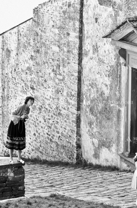 A woman standing on a little wall with a camera in her hands. Beside her, the entrance of a building. Fiesole, 1950s SPECIAL FEE  - CONTACT US FOR NOT EDITORIAL USAGE
