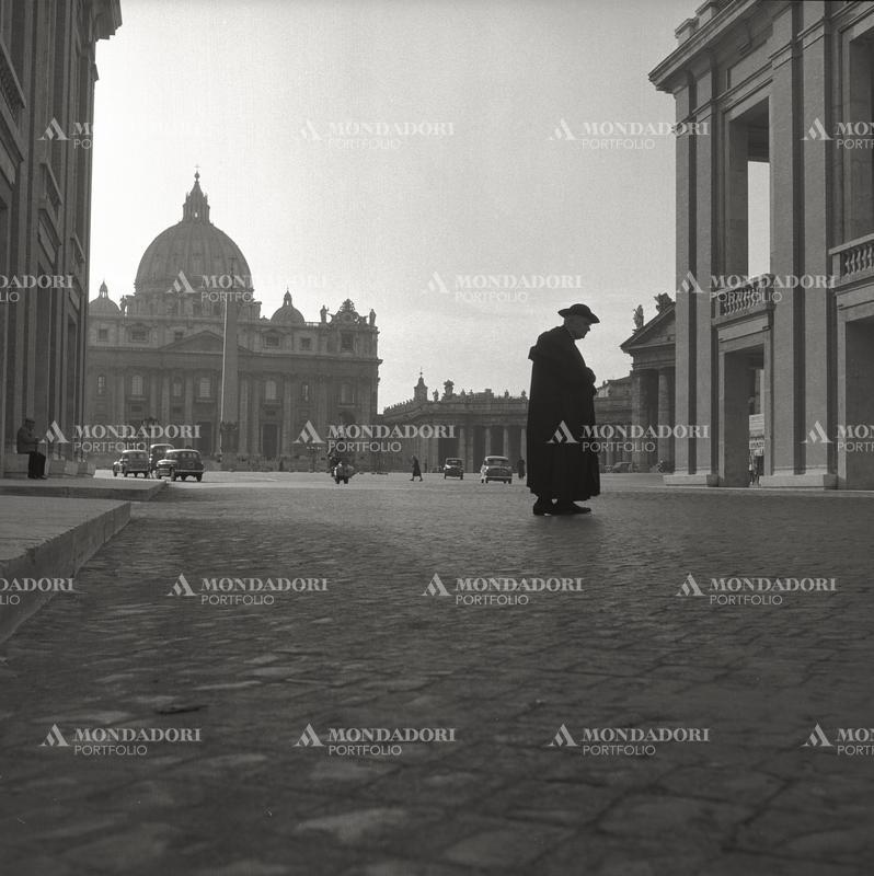 "An old priest crosses a street; in the background it is possible to see Papal Basilica of Saint Peter and its colonnade. Rome, 1955." SPECIAL FEE  - CONTACT US FOR NOT EDITORIAL USAGE