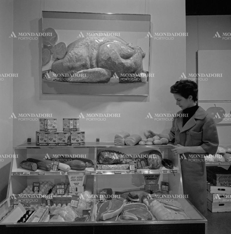 A woman observing the installation The American Supermarket by Robert Watts displayed at Il segno art gallery for the exhibition American Supermarket. A painting depicting a tridimensional turkey painted by Tom Wesselmann hanged above a shelf with Del Monte prunes, charcuterie and eggs. Rome, March 1965 SPECIAL FEE  - CONTACT US FOR NOT EDITORIAL USAGE