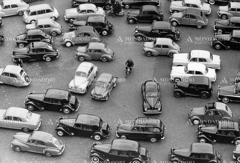 View from above of a street crowded with cars lying around; in the middle, a man by motor scooter passes the space through a car and another. Paris (France), April 1954. SPECIAL FEE  - CONTACT US FOR NOT EDITORIAL USAGE