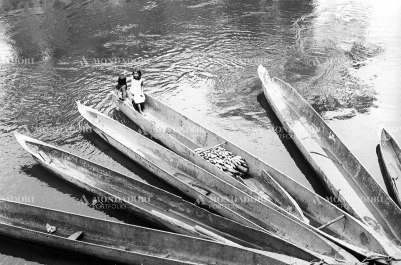 Two girls on a canoe on which are loaded some bananas, among other canoes. Latin America, 1970. SPECIAL FEE  - CONTACT US FOR NOT EDITORIAL USAGE